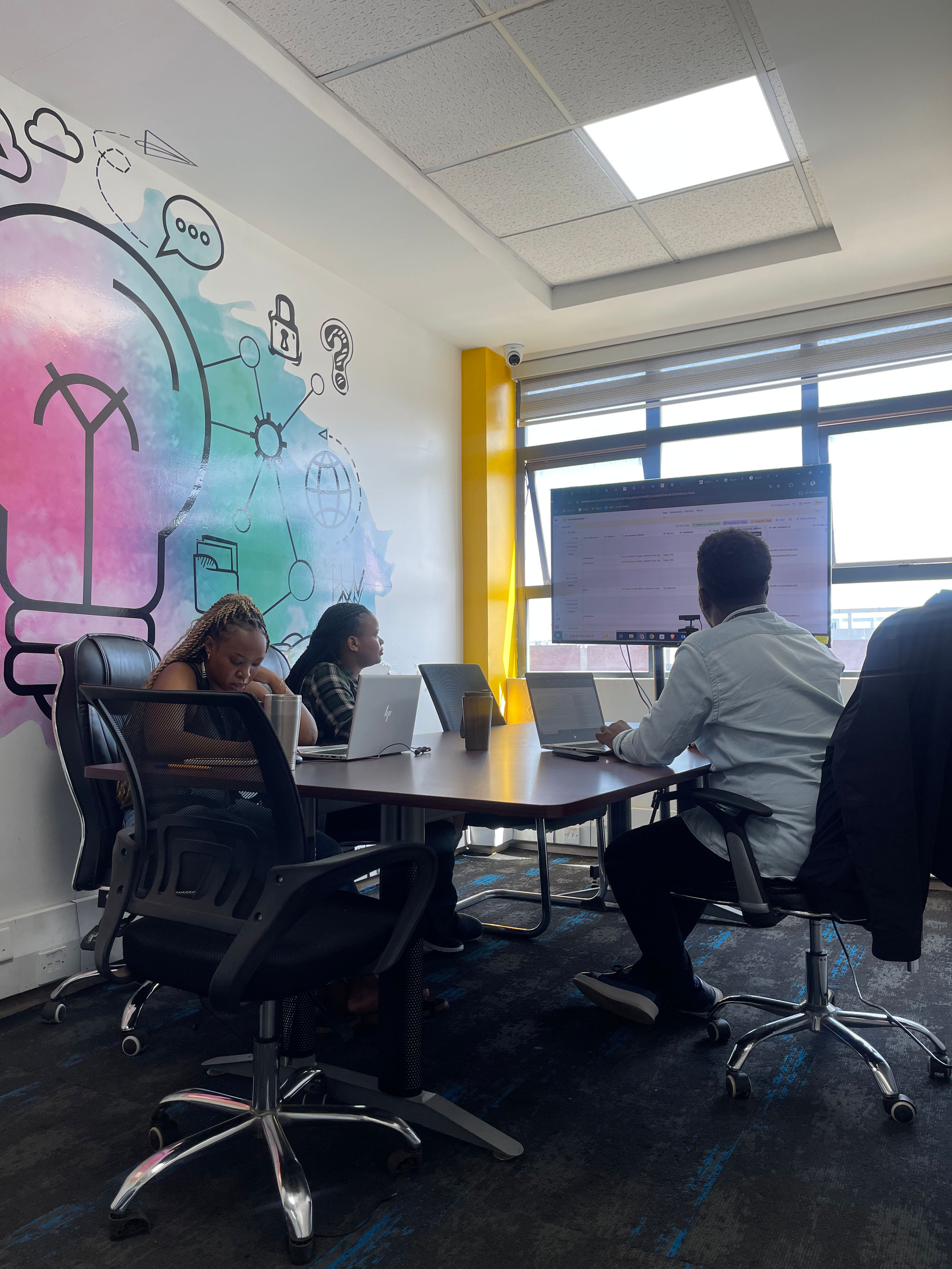 Impact Outsourcing QA reviewers working at a shared table, laptops open alongside a wall-mounted review monitor.