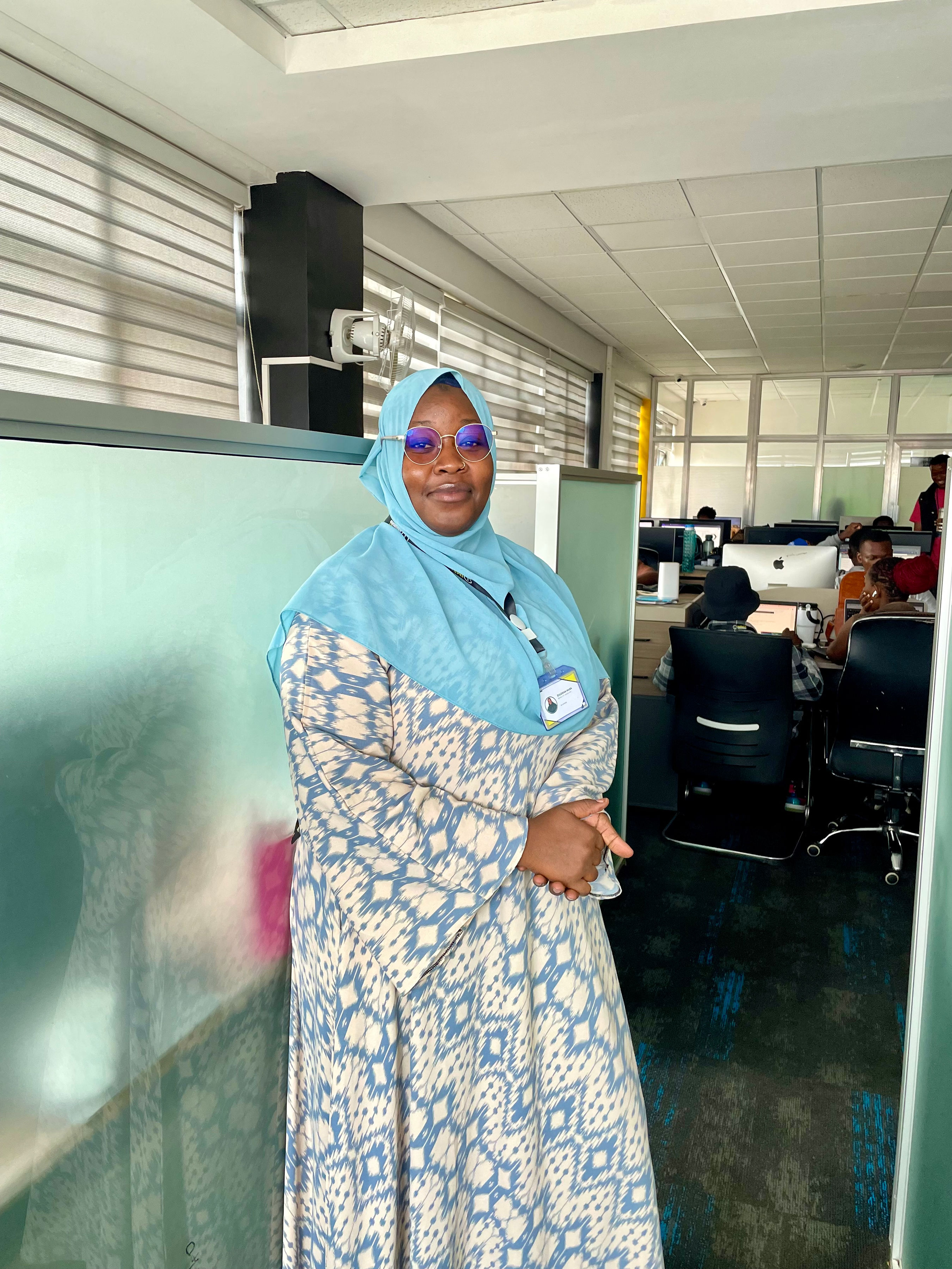 An Impact Outsourcing delivery team member on the Nairobi floor, with the wider workstation bay visible behind her.