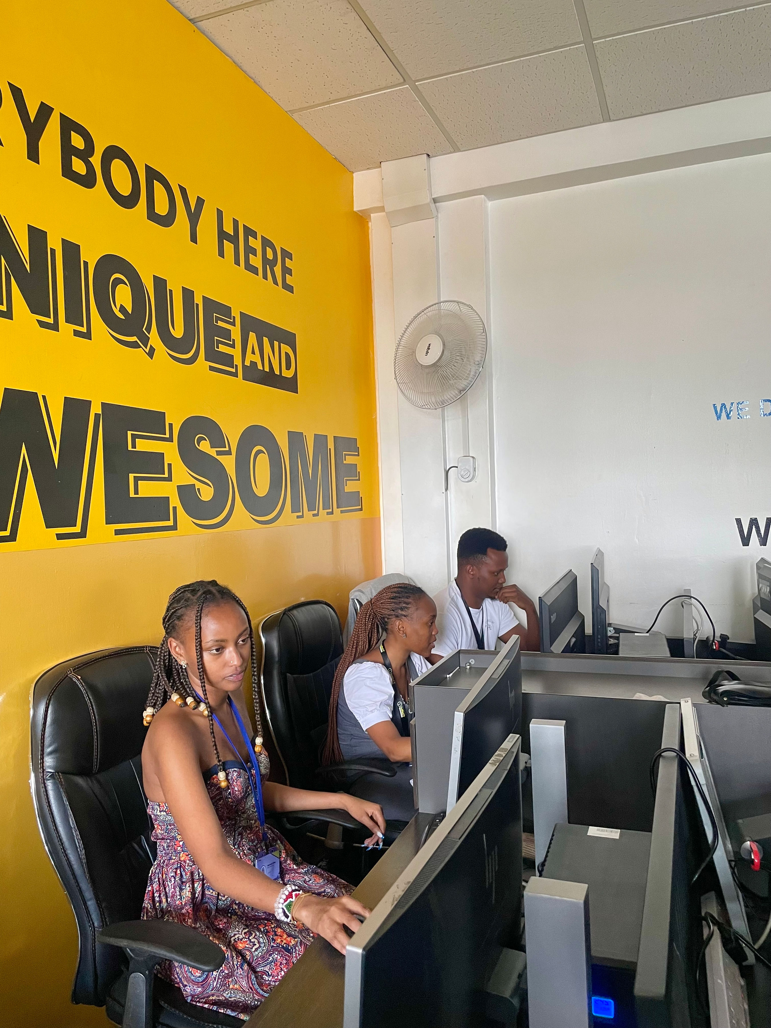 Impact Outsourcing operators at their workstations in the Nairobi delivery floor, with an 'Everybody here is unique and awesome' mural visible behind them.